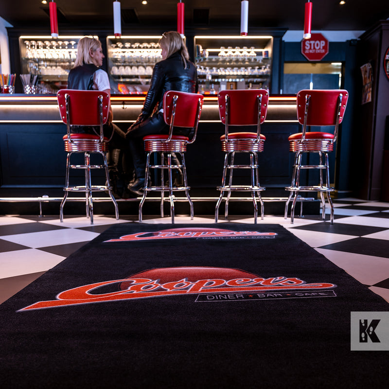 Bar scene with two ladies sat on stools and a large branded floor mat