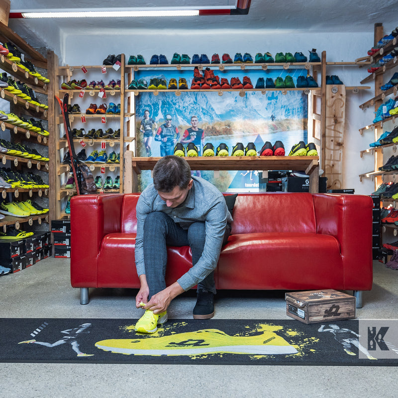 A person sitting on a red couch is trying shoes on a black mat with a realistic print, placed on a floor with various shoes displayed on shelves in the background.