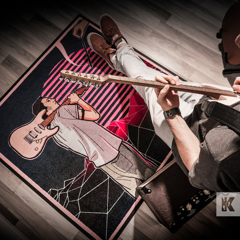 Man plays guitar while sat on an amplifier with an album cover design on a printed mat
