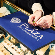 A branded counter mat with the text 'PLAZA 20th Century Jewellery' being used as a work surface by a person handling jewelry items.