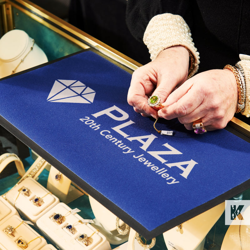 A branded counter mat with the text 'PLAZA 20th Century Jewellery' being used as a work surface by a person handling jewelry items.