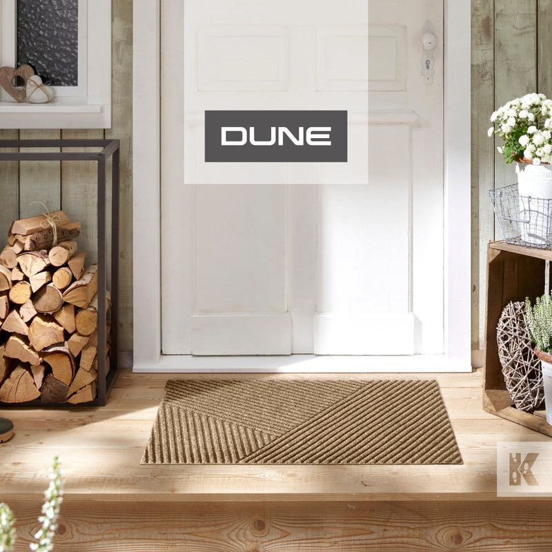A beige indoor scraper mat with a geometric pattern, placed on a wooden floor in front of a white door, with the brand name 'DUNE' visible.