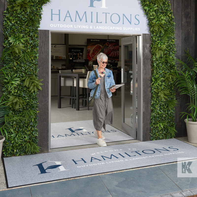 A custom logo mat printed with the name 'Hamiltons' entrance mat displayed outside a business establishment with a woman standing beside it.
