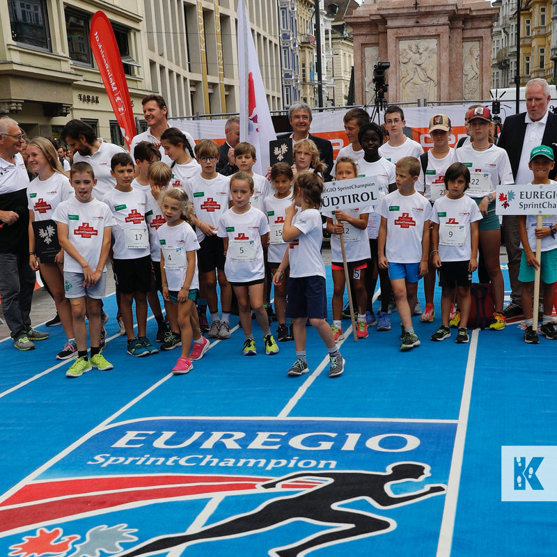 A group of children and adults standing on a blue running track with a banner that reads 'Eurogio Sporting Club' and 'Eurogio Spirit Champion'. There are also signs with the word 'UDTIROL' and the logo of the event.