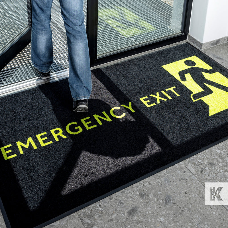 A black door mat with the words 'EMERGENCY EXIT' printed in yellow, placed on a door threshold with a person's feet visible stepping onto it.