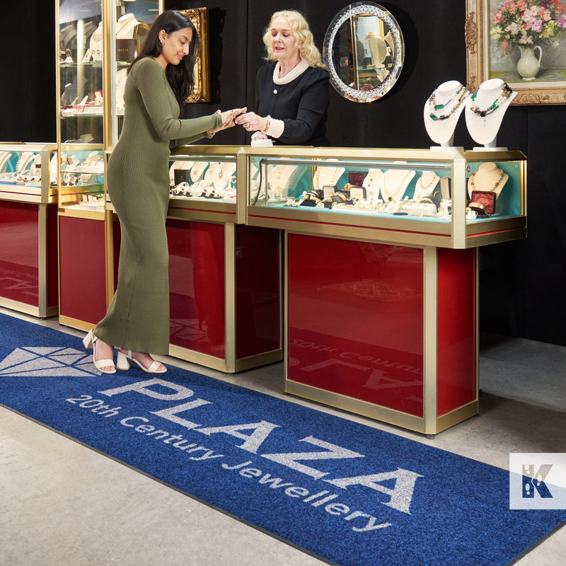 A printed advertisement mat with the text 'Plaza 20th Century Jewellery' displayed at a jewellery store counter.