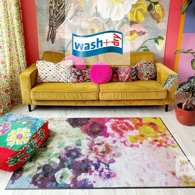 A colorful floral designer floormat laid out on a floor with a yellow couch and various pillows in the background, in a room with vibrant wall decorations.