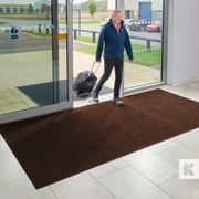 Man with a suitcase exiting a building onto a brown doormat.